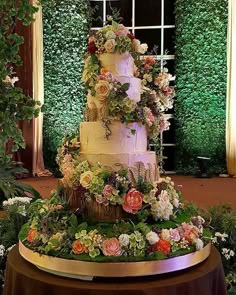 a three tiered wedding cake is surrounded by greenery and flowers in front of a window