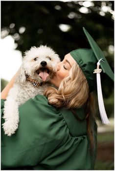 a woman in a green graduation gown kissing a white dog on the cheek while wearing a cap and gown