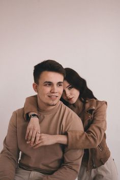 a man and woman sitting next to each other in front of a white wall with their arms around each other