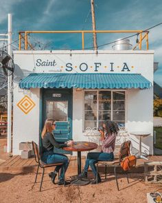 two women sitting at a table in front of a small store with blue awnings