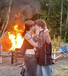 two people standing next to each other in front of a fire with their arms around one another