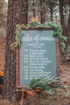 a chalkboard sign with greenery on it next to a tree in the woods