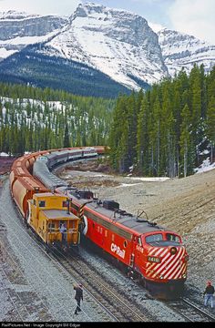 a red and yellow train traveling down tracks next to snow covered mountain range with evergreen trees