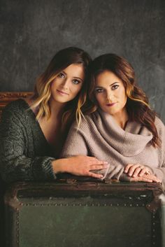 two beautiful women sitting next to each other in front of an old suitcase and posing for the camera