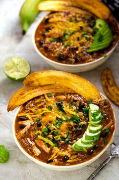 two white bowls filled with chili, beans and tortilla chips next to lime wedges