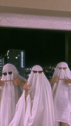 four women dressed in white veils are standing near a window with city lights behind them