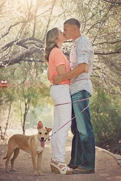 a man and woman kissing while standing next to a dog on a leash with trees in the background