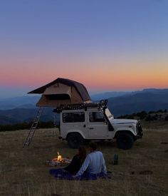 two people sitting in the grass under an awning next to a camper van