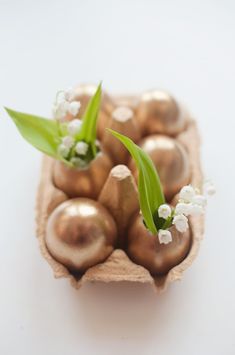 small gold eggs in a carton with flowers on the top and green stems sticking out of them