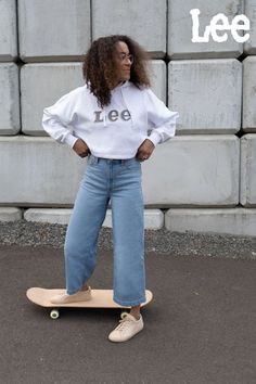 Woman outdoors standing in front of a cement wall on a dark sidewalk with a tan skateboard under her right foot. Her left foot is standing on the sidewalk as she is posing for the camera with her hands on her hips. She is wearing tan simple sneakers, high rise wide leg cropped jeans and a white Lee logo cropped sweatshirt. She is looking over her left shoulder away from the camera. Comfort Vibes, Comfy Winter Outfits, Cropped Sweatshirt, Crop Jeans, White Hoodie