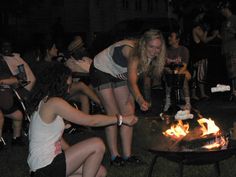 people sitting around a fire pit at night with one person lighting the flame and another standing in front of it