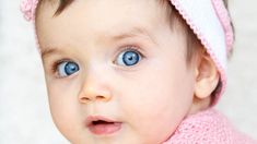 a close up of a baby wearing a pink sweater and a white hat with blue eyes