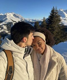 a man and woman standing next to each other in the snow with mountains behind them