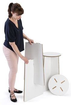 a woman standing next to a white table with two pieces of cardboard on it and another piece of furniture in front of her