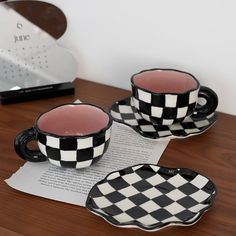 two black and white checkered coffee cups sitting on top of a wooden table next to an open book