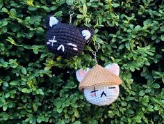 two crocheted cats hanging from chains on top of green bushes, one is wearing a hat and the other has eyes