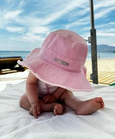 a baby wearing a pink hat sitting on top of a white blanket next to the ocean