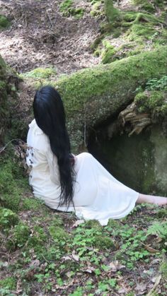 a woman with long black hair sitting on the ground in front of a mossy rock