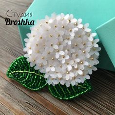 a brooch with white flowers and green leaves on a wooden table next to a blue box