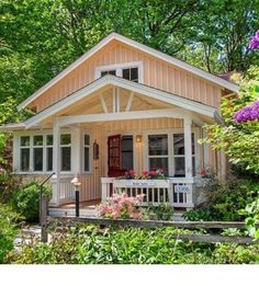 a small white house surrounded by trees and flowers in front of the door is a porch