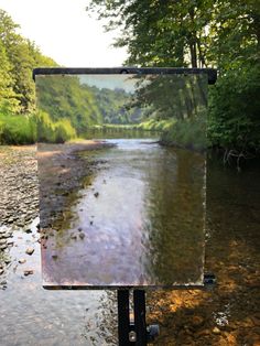 an easel with a painting on it sitting in the middle of a river surrounded by trees