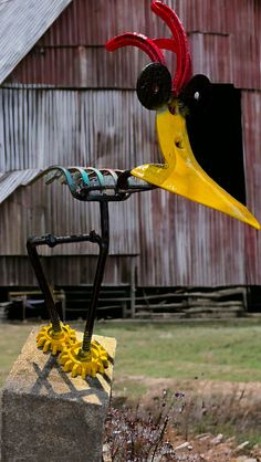 a yellow and red bird sculpture sitting on top of a cement block next to a barn