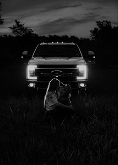 a woman sitting in the grass next to a truck at night with her dog on her lap