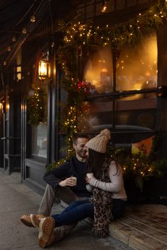 a man and woman are sitting on the steps in front of a building with christmas lights