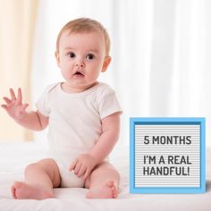 a baby sitting on top of a bed next to a sign that says 5 months i'm a real handful