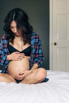 a pregnant woman sitting on top of a bed wearing a black bra and plaid shirt