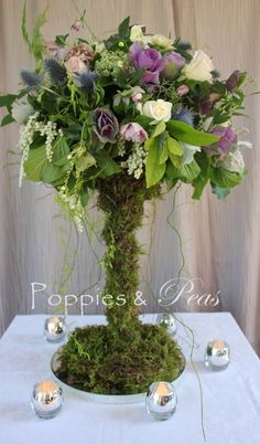 an arrangement of flowers and greenery in a vase on a white tablecloth with candles