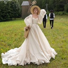 a woman in a white dress and hat walking across a grass covered field next to a man