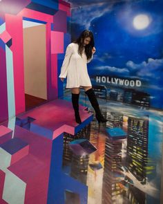 a woman is standing in front of a hollywood sign with buildings painted on the wall