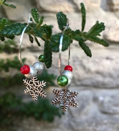 two silver snowflake earrings hanging from a branch with green and red beads on them