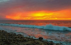 the sun is setting over the ocean with waves crashing in front of it and rocks on the shore