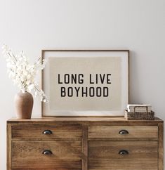 a wooden dresser topped with a vase filled with flowers next to a framed poster that says long live boyhood