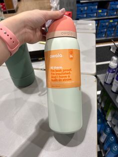 a hand is holding an orange and white water bottle in front of shelves with bottles