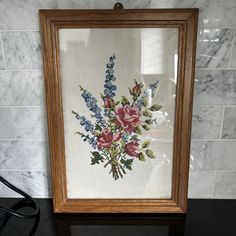 a cross stitch floral arrangement in a wooden frame on a marble tiled wall next to a black table