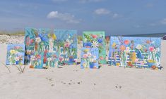 four paintings are displayed on the beach in front of an ocean and blue sky with white clouds