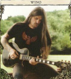 a man with long hair playing an electric guitar in front of a green field and trees