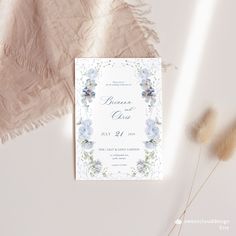 a white and blue wedding card with flowers on it next to some pamy grass