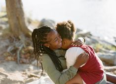 two children hugging each other while sitting on the ground next to a tree and water