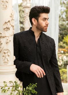 a man standing next to a white pillar wearing a black shirt and pants with an embellished design on it