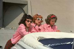 three women in pink jackets and sunglasses sitting on the back of a car with their arms around each other