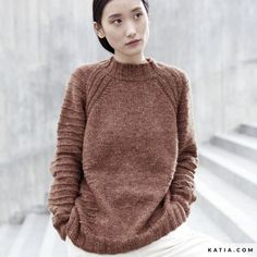 a woman standing in front of some stairs wearing a brown sweater and white pants with her hands on her hips