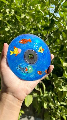 a hand holding a blue frisbee in front of some bushes and trees with fish painted on it