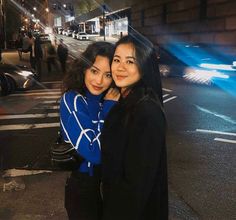 two women standing next to each other in the street at night time, one holding her arm around another woman's shoulder