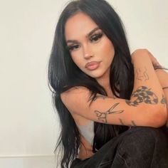 a woman with long black hair and tattoos on her arm sitting next to a wall