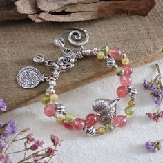 This artisan handmade double-strand bracelet showcases the natural beauty of beautiful grade gemstones, including Strawberry Quartz (10.8mm), Rutilated Quartz (6.8 mm), and Prehnite (8 mm), that carefully selected to make the colors of this bracelet stand out, creating a harmonious blend of pink, green, and yellow hues. The bracelet is adorned with intricate Karen Hill Tribe fine silver accents, featuring charming elements like a hammered heart-shaped bead, flower S-clasp, and unique charms, Including 925 sterling silver, all expertly strung together with a strong and durable waxed cord, finished with woven ends and Hill Tribe fine silver chain. Whether you're looking to add a statement piece to your jewelry collection, attend a festival or a boho-themed event, search for a thoughtful gift Bohemian Tourmaline Bead Jewelry, Bohemian Tourmaline Round Beads Jewelry, Spiritual Tourmaline Beaded Bracelets With Natural Stones, Bohemian Beaded Tourmaline Jewelry, Bohemian Tourmaline Jewelry With Natural Stones, Tourmaline Gemstone Beads Bracelets For Jewelry Making, Multi-strand Natural Stone Bracelets As Gift, Multi-strand Natural Stones Bracelet For Gifts, Multi-strand Natural Stone Bracelets For Gifts