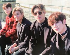 four young men sitting next to each other in front of some water and one is wearing sunglasses